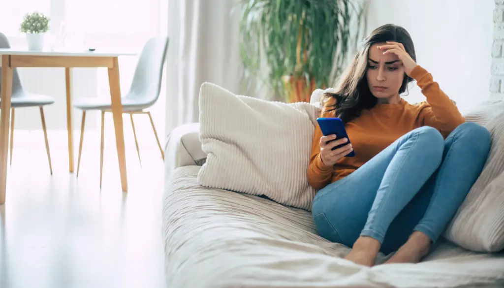 A woman on a sofa, reading her phone and looking worried.