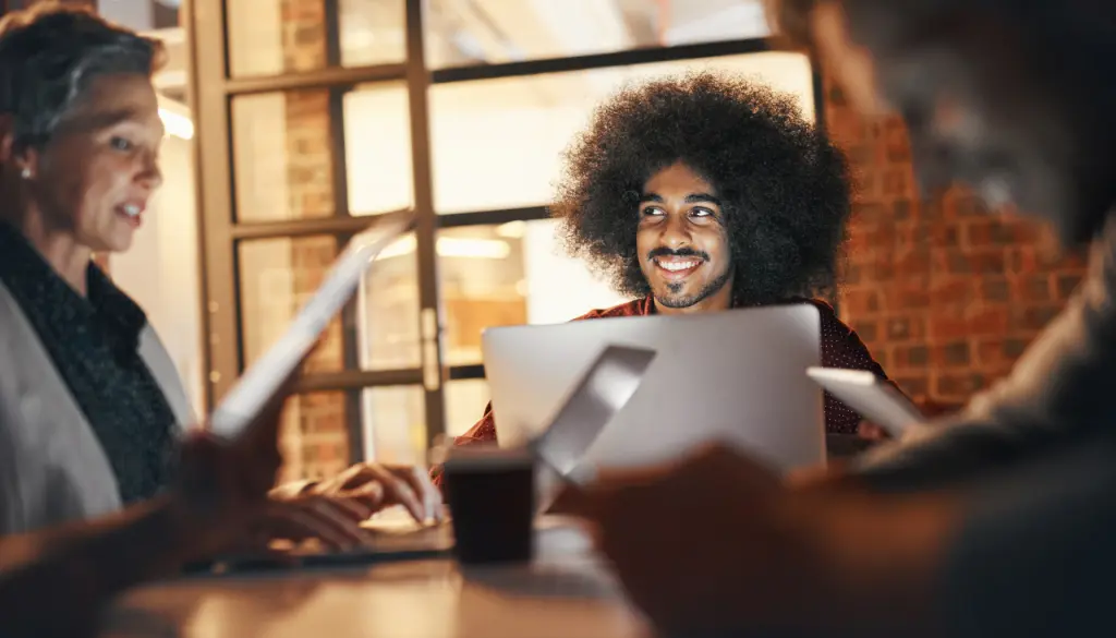 A man smiling as he talks with others around a laptop.
