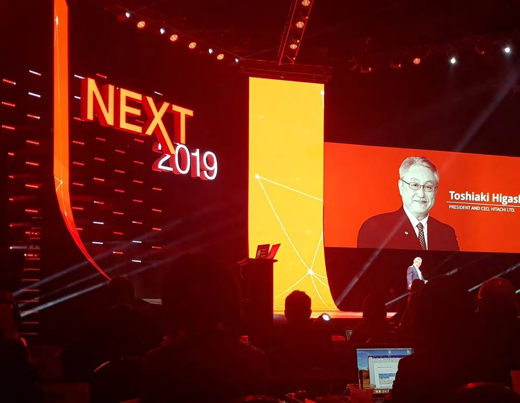 The keynote stage at Hitachi NEXT 2019: the NEXT 2019 sign, and Hitachi Ltd.'s president and CEO on the big screen above the audience.