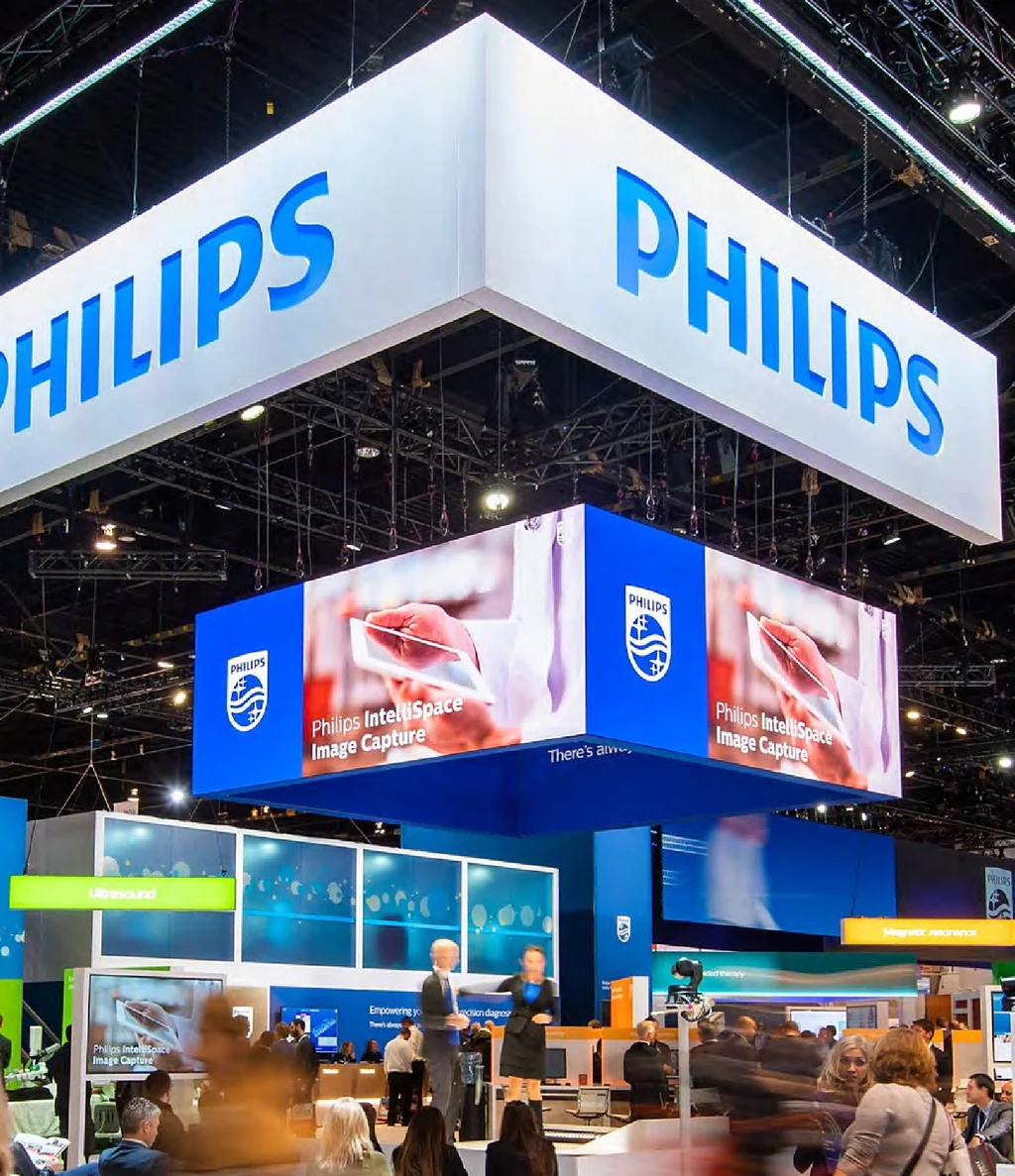 A busy Philips exhibition stand under a large hanging Philips sign and two wide screens.