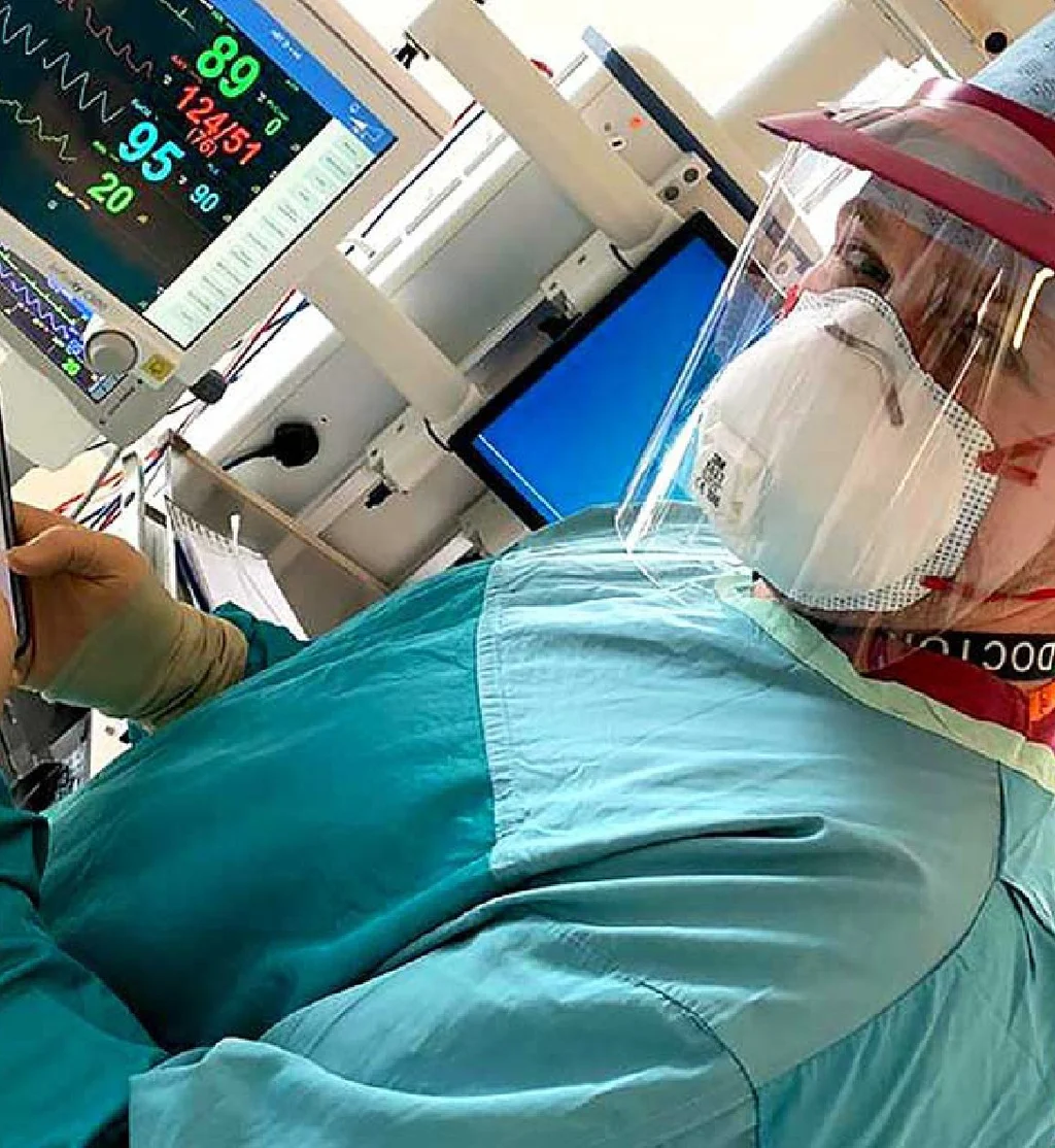A doctor in scrubs, a face shield and a mask at a patient monitor, a phone in a gloved hand.